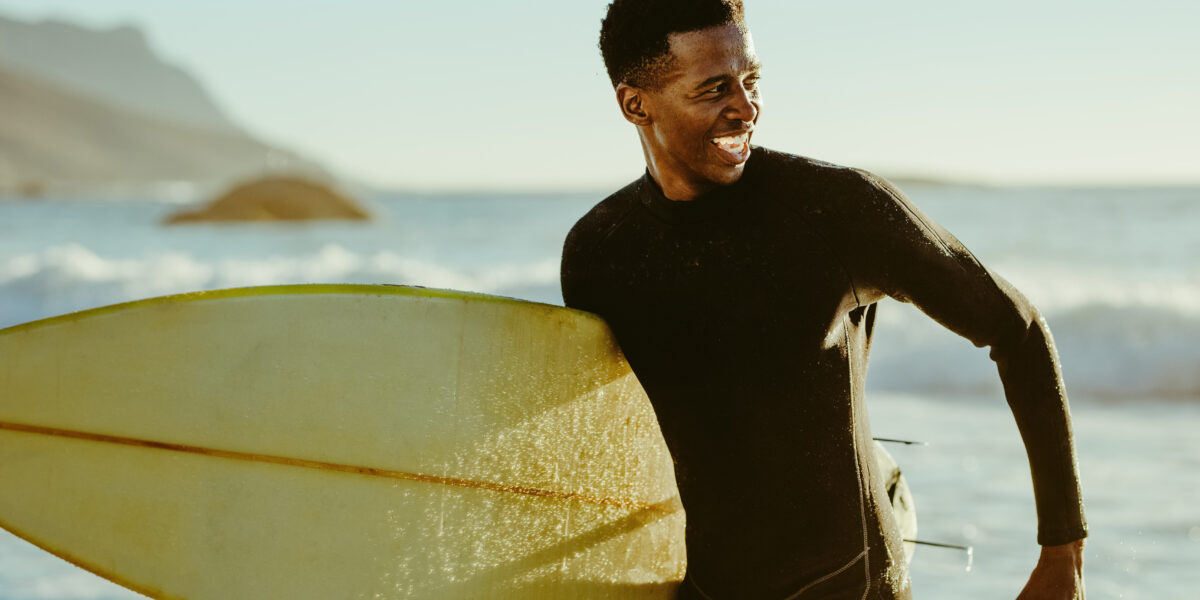 Smiling african male running out of the ocean after water surfing. Happy young man with surfboard on the beach enjoying holidays.