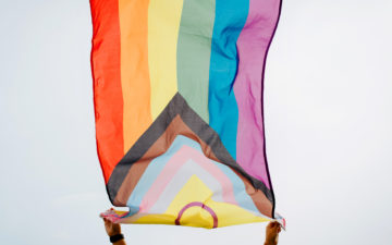 Pride 2022: Rainbow flag, new international symbol for LGBTQ + community, gay, lesbian, transperson and people of color. Person holding waving LGBT flag against clear sky background, low angle view