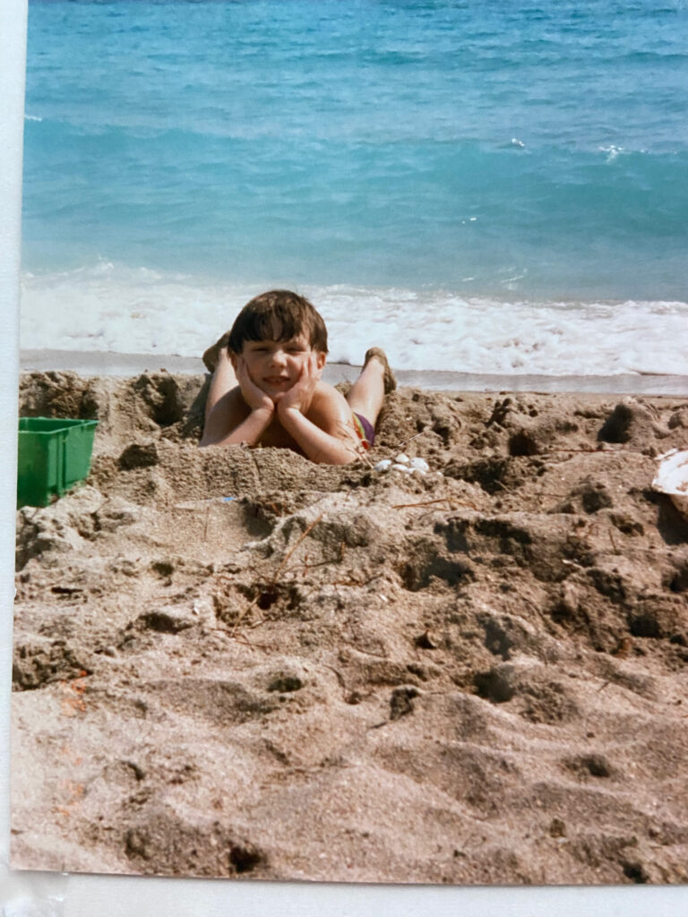 Ben as a child laying in the sand and smiling, with a green bucket next to him