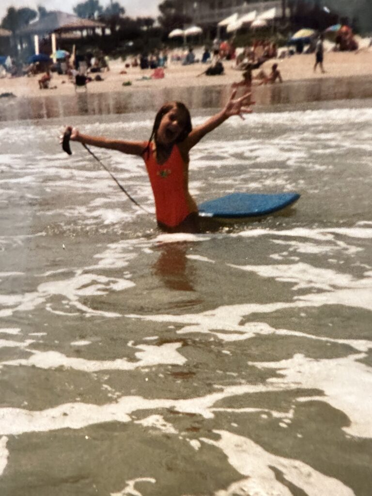 Alex Refosco as a child with her blue boogey board, throwing her hands up in the air while standing in the ocean