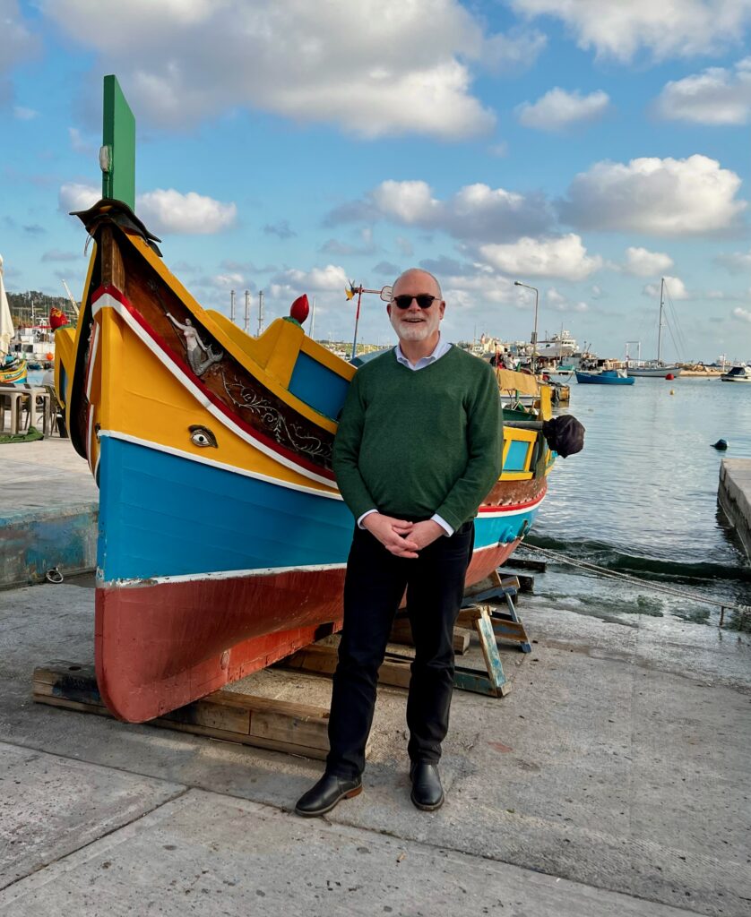 Mark J. Spalding se tient devant un bateau coloré à Malte.