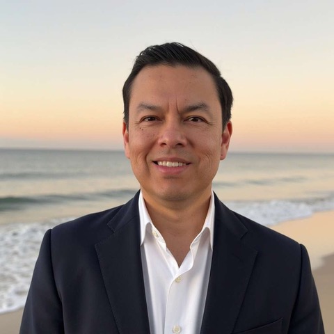 Rolando Morillo dressed in a white button up shirt and black blaze with the ocean behind him.